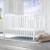 White crib with a teddy bear on a wooden floor in a bright room with large windows. 16-Taylor 4-in-1 Convertible Crib