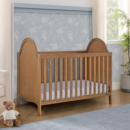 Wooden crib in a nursery with floral wallpaper and a teddy bear.