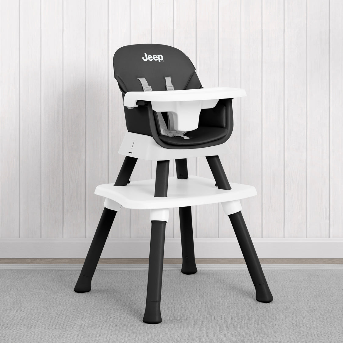 A black and white Jeep high chair with a white tray and seat, set against a white paneled wall and gray carpet.