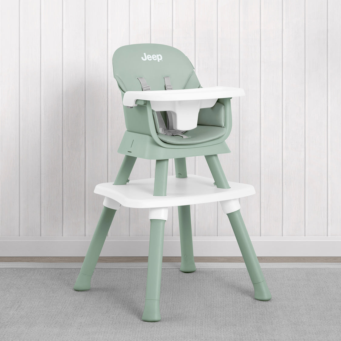 A light sage green and white plastic high chair with a matching footrest, shown against a white wood-paneled wall and gray carpet. The brand name 'Jeep' is printed on the backrest.