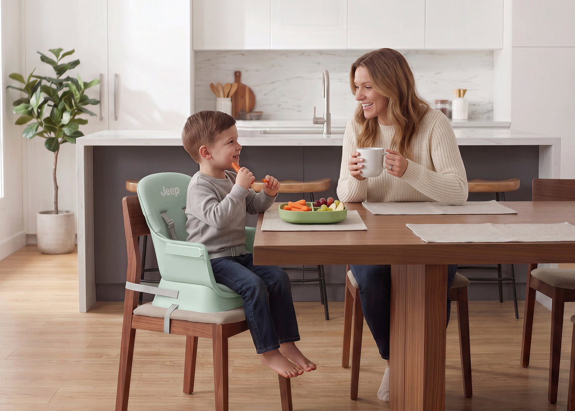 A woman and a young boy share a meal at a wooden dining table in a bright, modern kitchen. The boy is seated in a light mint green Jero toddler booster seat attached to a wooden chair, while the woman holds a white mug.