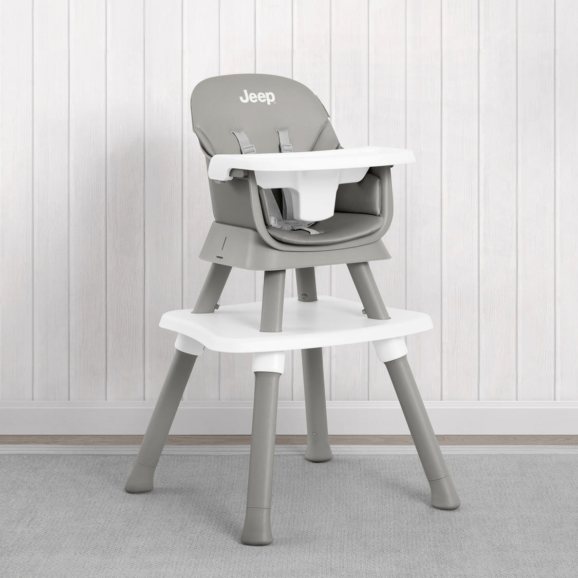 A gray and white Jeep high chair with a matching footrest, set against a white paneled wall and gray carpet.