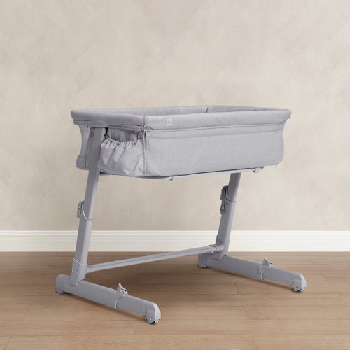 A light gray adjustable bassinet on a matching gray support stand, set against a neutral beige textured wall and light wood floor.