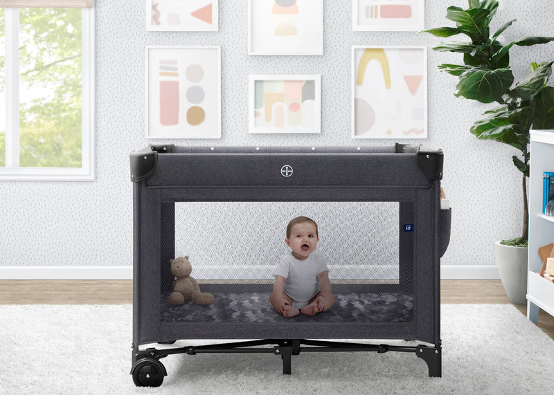 Baby sitting inside a portable crib in a room with a window, plant, and framed pictures on the wall.