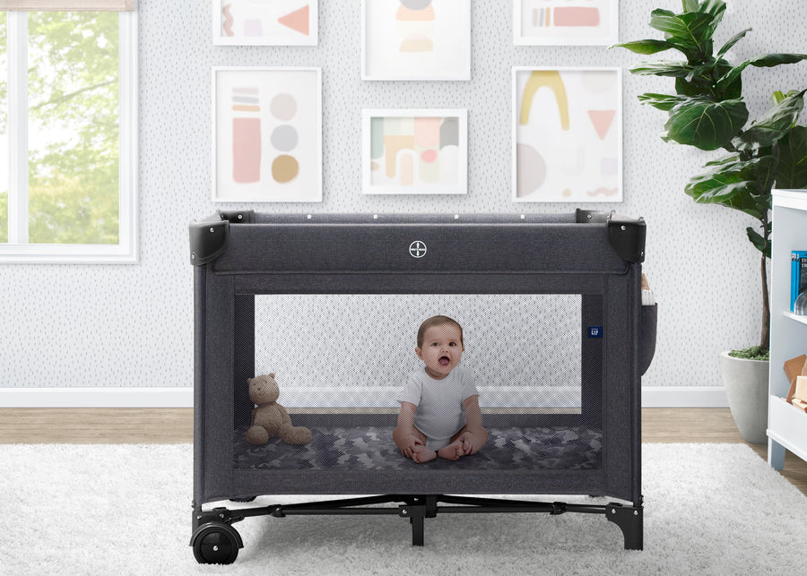 Baby sitting inside a portable crib in a room with framed art on the wall. 11-babyGap Deluxe Play Yard