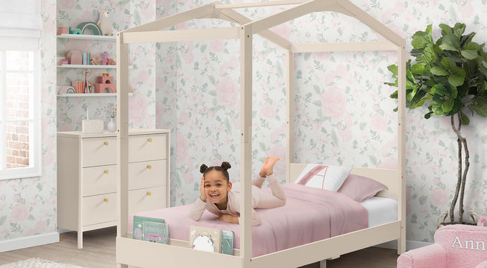 Child in a house-shaped bed with pink bedding in a room with floral wallpaper and a dresser. 0-Poppy Collection