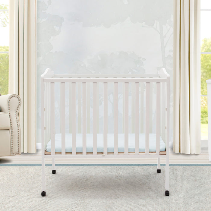 White crib in a bright nursery with large windows and curtains. 3-Folding Portable Mini Baby Crib with 1.5-inch Mattress