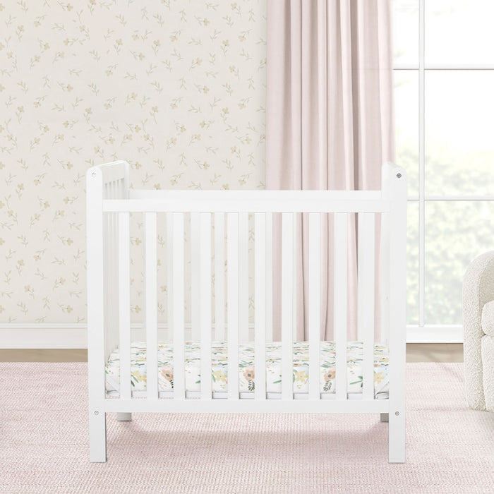 White crib in a nursery with floral wallpaper and pink curtains 2-Classic Mini Crib with 2.75-inch Mattress - Convertible to Twin Bed