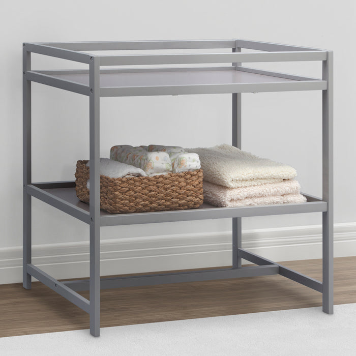 A gray Harbor Changing Table from Delta Children with open shelves, dressed with folded baby clothes and a basket. 3-Harbor Changing Table
