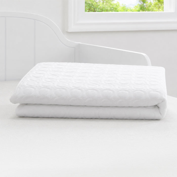 A folded white quilted mattress topper or pillowcase sits on a white bed in a bright, minimalist bedroom with a window and white headboard. 3-Eco-Cool Cooling Crib Mattress Protector