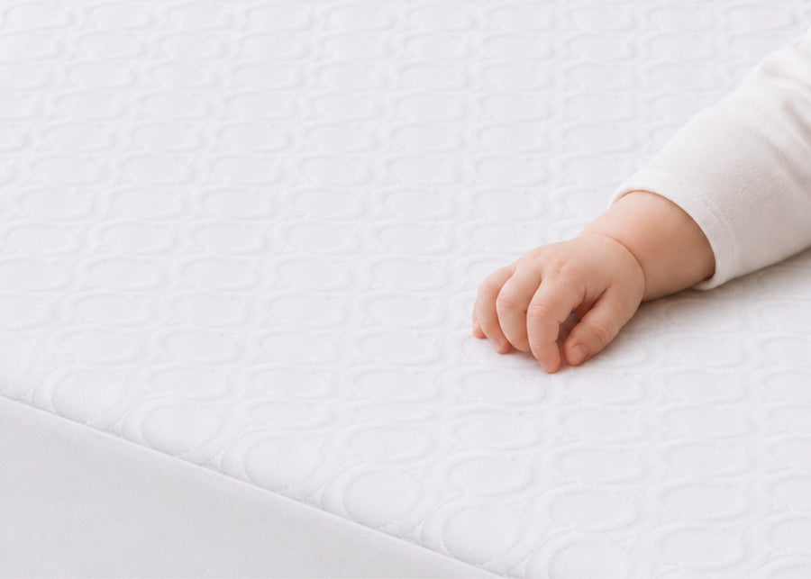 A close-up of a baby's hand resting on a white, quilted mattress topper or mattress surface. 9-Eco-Cool Cooling Crib Mattress Protector