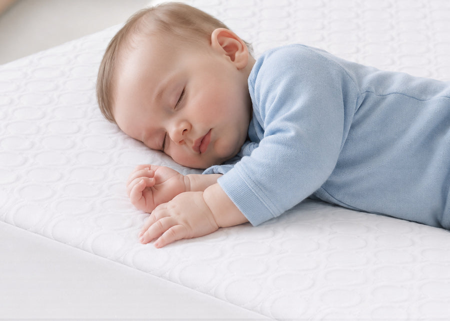 A sleeping baby wearing a light blue long-sleeve onesie, lying on a white textured mattress. 14-Eco-Cool Cooling Crib Mattress Protector