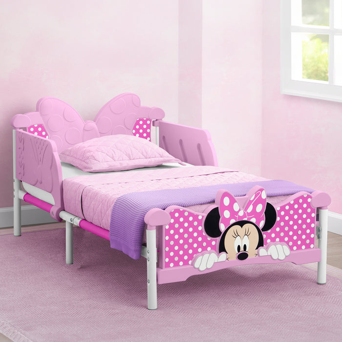 A toddler-sized bed with a Minnie Mouse theme, featuring a headboard, footboard, and side rails with Minnie Mouse decals and three-dimensional accents, placed in a room with a pink carpet and decorative items. 0-Minnie Mouse 3D Toddler Bed