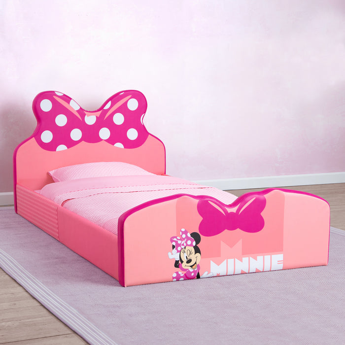 A pink upholstered twin bed with a bow detail and Minnie Mouse character decorations in a children's bedroom. 1-Minnie Mouse Upholstered Twin Bed