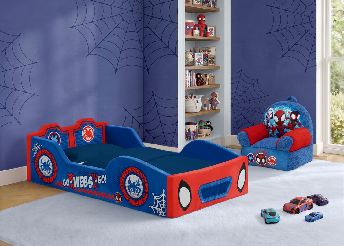 A children's bed styled with Spider-Man graphics, featuring a blue and red frame with spider web patterns and character decals. The bed is shown in a decorated bedroom with Spider-Man themed wallpaper, a matching armchair, and toy cars on the floor.