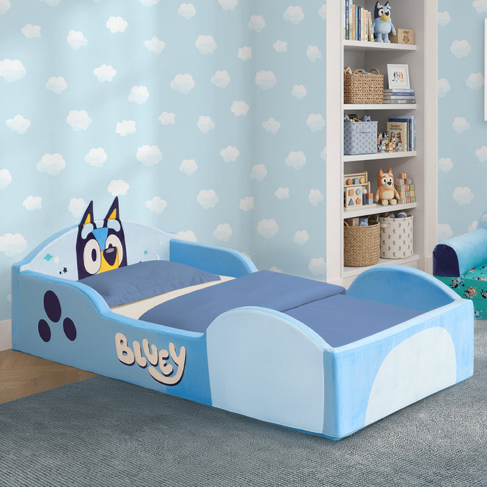 A children's bed styled like Bluey, featuring a light blue and white frame with the character's face on the headboard, blue bedding, and a matching blue patterned wall in a child's bedroom. 0-Bluey Foam Frame Toddler Bed