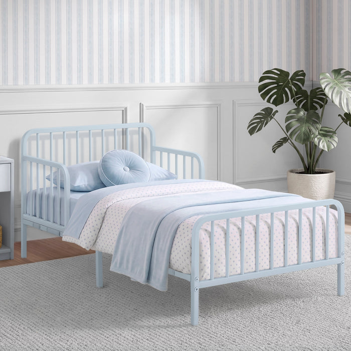 A light blue metal toddler bed with a guard rail, styled with a light blue and white patterned duvet, matching pillow, and a round tufted pillow, set in a child's bedroom with striped wallpaper and a potted plant. 3-Finley Metal Toddler Bed with Spindles
