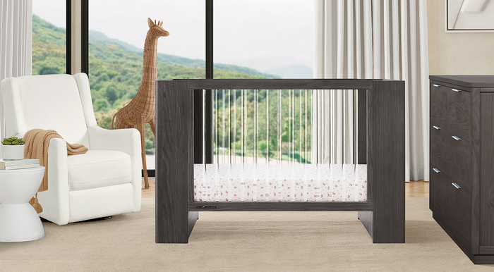 A modern white nursery set including a crib with a white guardrail, a white glider chair, a dresser, and a changing top, set in a room with a large window and a stuffed animal on the chair. 0-Aerin Collection