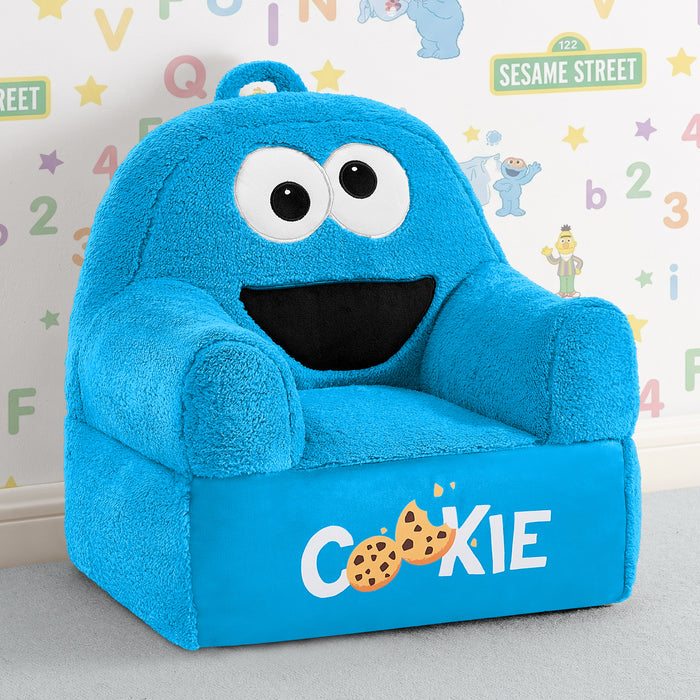 A plush blue children's armchair designed like the Cookie Monster from Sesame Street, with large white eyes, a black mouth, and 'COOKIE' printed on the front with cookie graphics. The chair is photographed in a styled room setting with a patterned wall behind it. 1-Sesame Street Cookie Monster Cozee Buddy Chair
