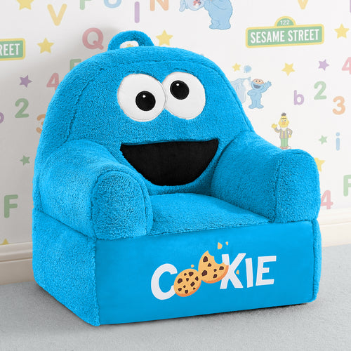 A plush blue children's armchair designed like the Cookie Monster from Sesame Street, with large white eyes, a black mouth, and 'COOKIE' printed on the front with cookie graphics. The chair is photographed in a styled room setting with a patterned wall behind it.