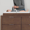 A baby changing table in dark brown wood with a white changing pad, shown with a baby and an adult standing behind it against a light gray wall. 9-EasyChange Changing Pad