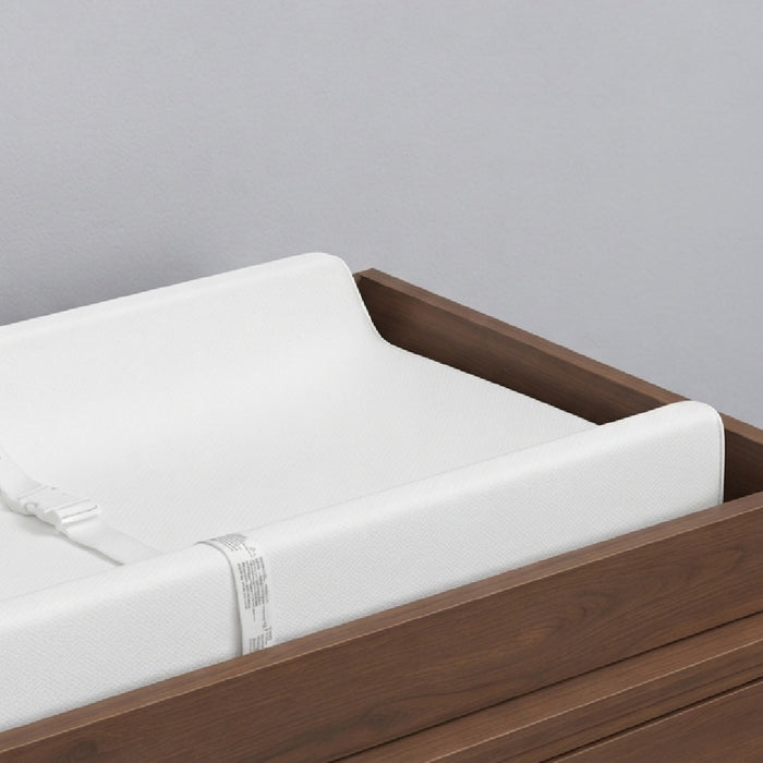 A close-up of a white changing pad with a strap, set inside a dark brown wooden changing table against a light gray background. 2-EasyChange Changing Pad