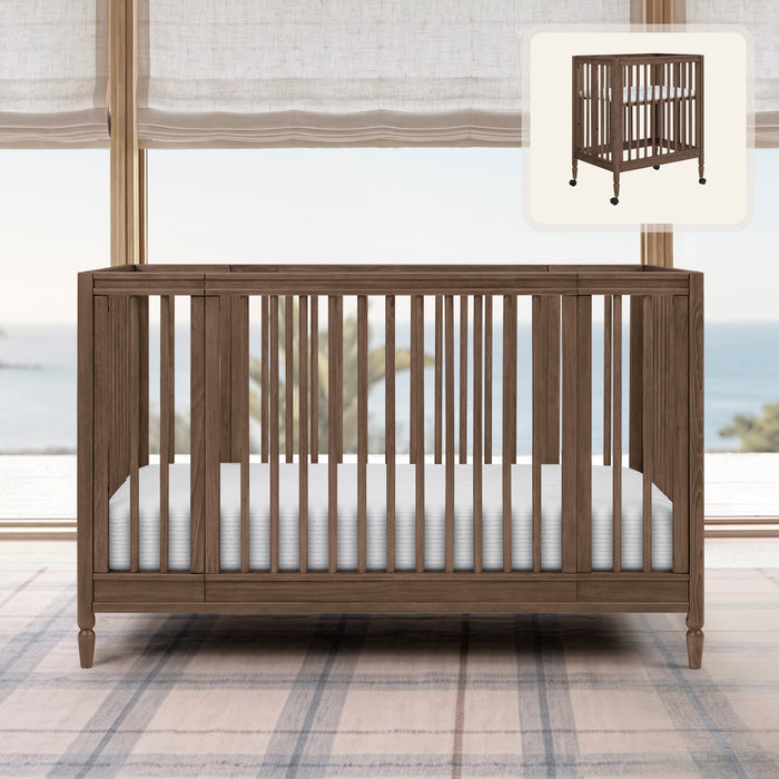 Wooden crib with white mattress in a room with plaid carpet and large windows. 0-Jalen Grow With Me 9-in-1 Convertible Bassinet to Crib