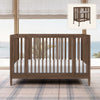 Wooden crib with white mattress in a room with plaid carpet and large windows. 25-Jalen Grow With Me 9-in-1 Convertible Bassinet to Crib