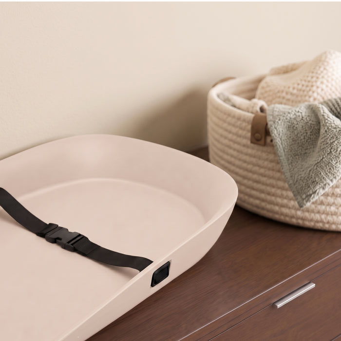 A cream-colored changing pad with a black safety strap resting on a dark wood dresser next to a woven basket. 0-Beautyrest Molded Waterproof Changing Pad with Fresh Technology