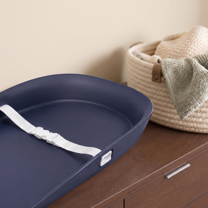 A navy blue changing pad with a white strap and buckle, styled on a dark wood dresser next to a beige woven basket. 1-Serta Molded Waterproof Changing Pad with Fresh Technology