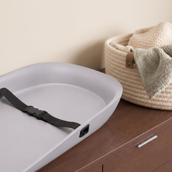 A light gray plastic changing pad tray with a black adjustable strap, shown on a dark wood dresser next to a woven basket. 2-Molded Waterproof Changing Pad with Fresh Technology