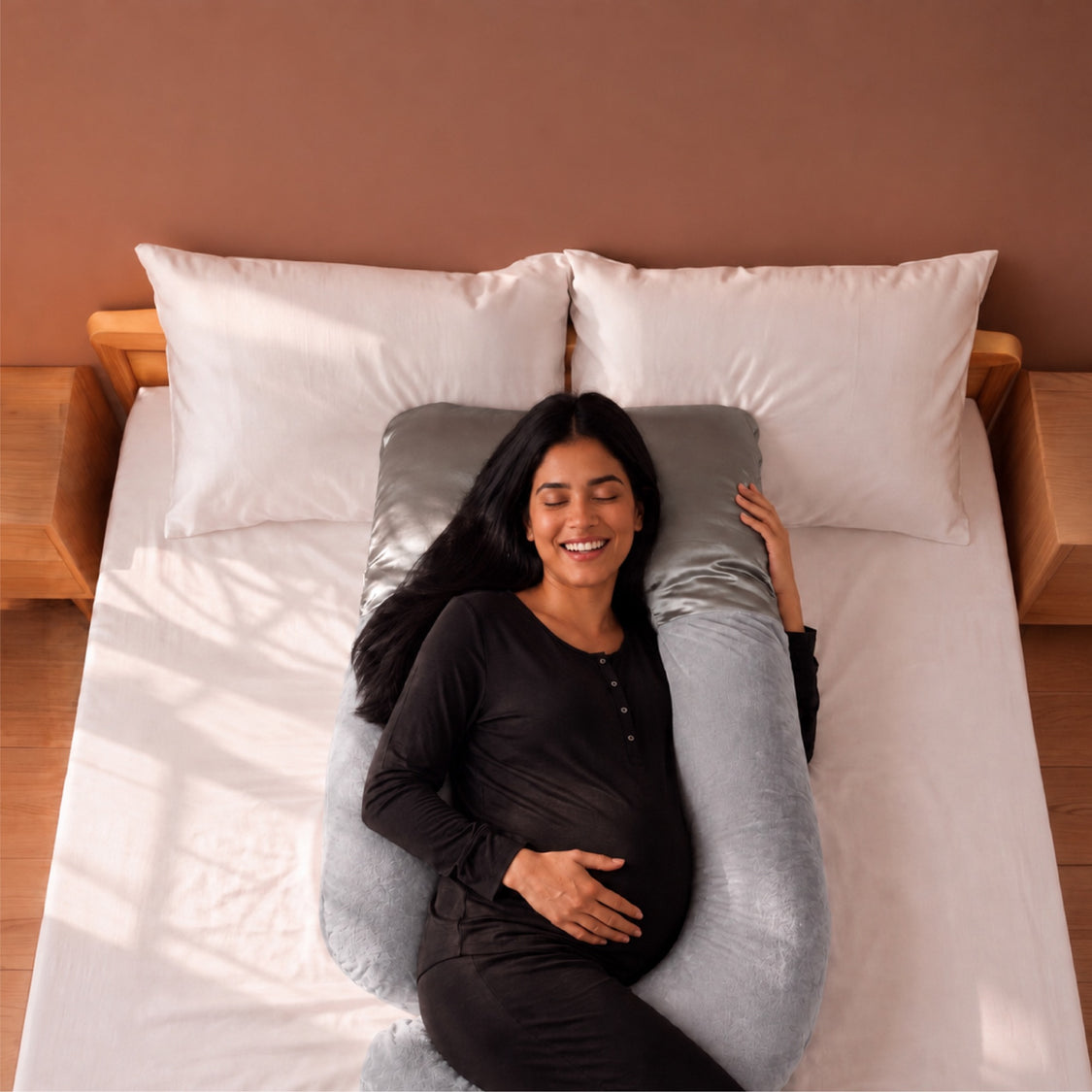A pregnant woman lies on a bed using a light gray U-shaped maternity pillow, with white pillows and a warm brown wall in the background.