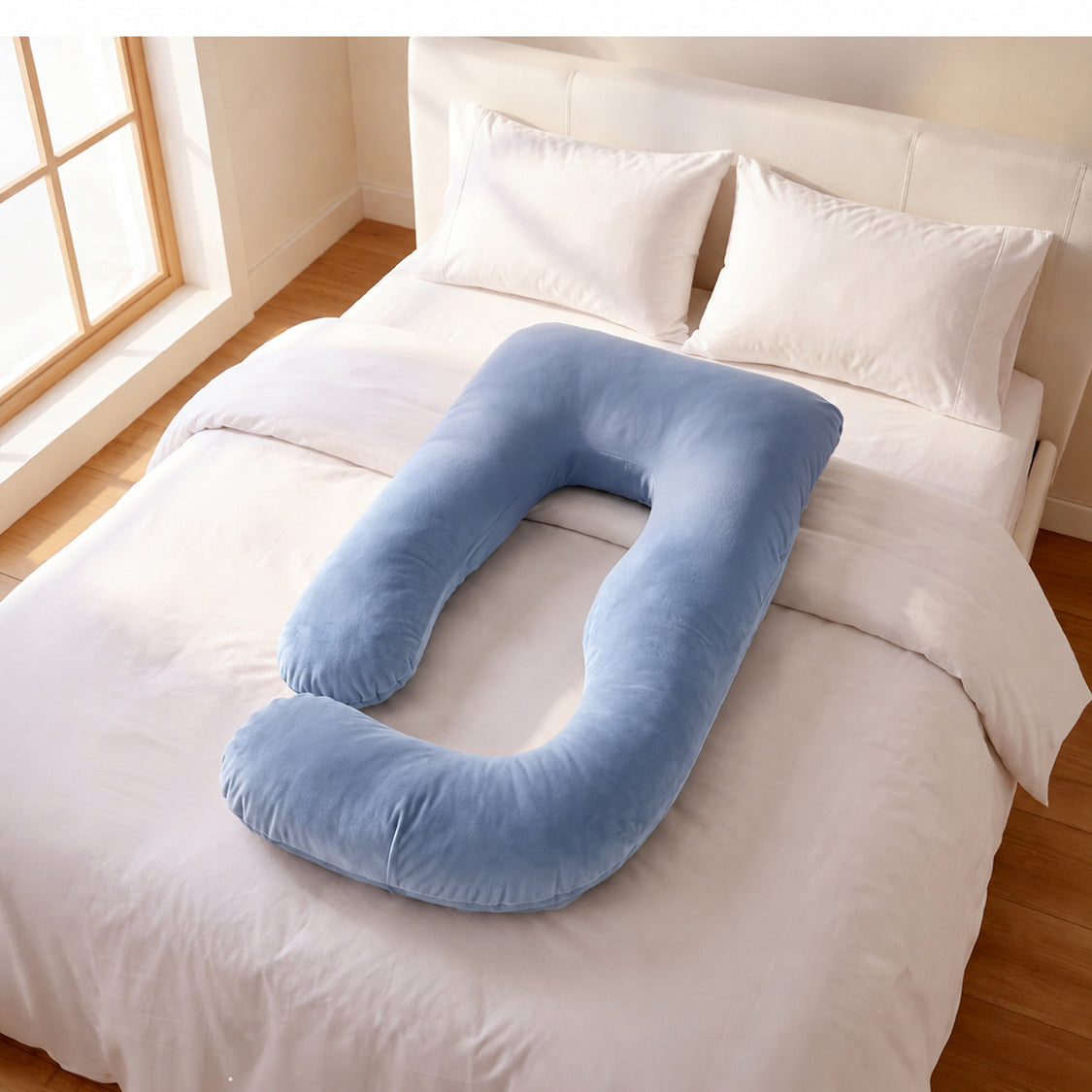 A light blue U-shaped nursing pillow resting on a neatly made bed with white linens and pillows in a bright bedroom.