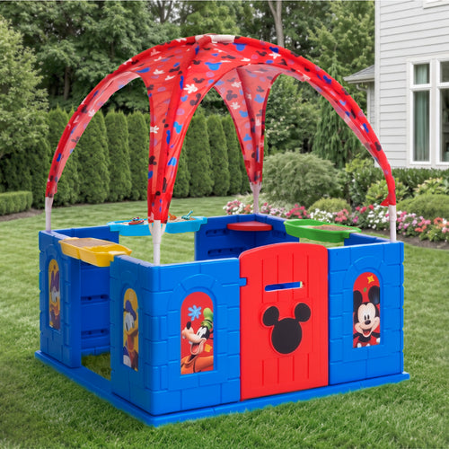A blue and red plastic children's play yard with a curved red canopy, set up on a green lawn in front of a house. The play yard features cartoon character windows and a red door with a black mouse head silhouette.