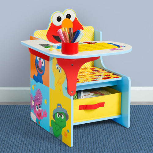 A colorful children's desk featuring Elmo on the backrest and cartoon character panels on the sides, with a red cup holding crayons on the desktop and a yellow storage drawer underneath, set against a light gray wall and blue carpet.