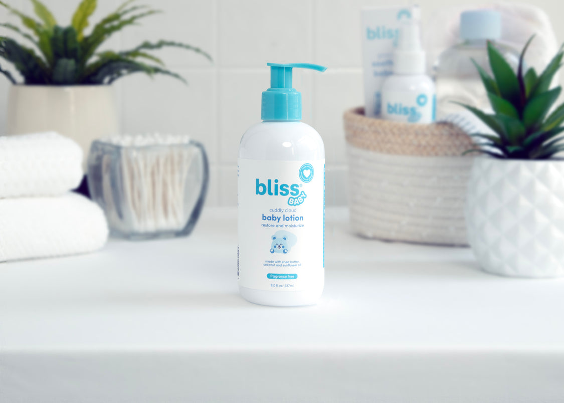 A bottle of Bliss baby lotion with a pump dispenser, placed on a bathroom shelf with decorative plants and bathroom essentials in the background.