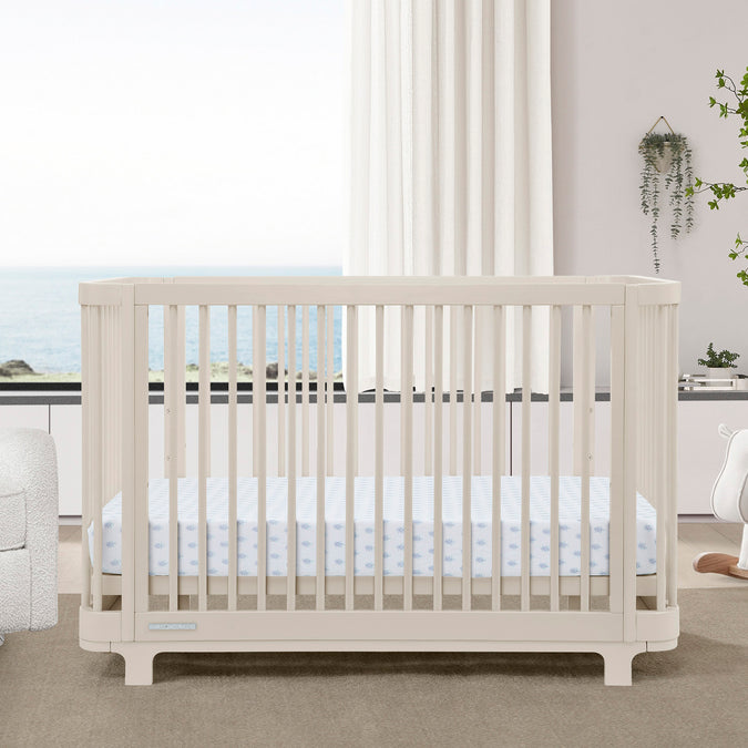A brown Nest 4-in-1 Convertible Crib by Delta Children in a modern nursery room, with a white mattress on a white floor, alongside a white chair and a wooden dresser. 65-Nest 4-in-1 Convertible Crib