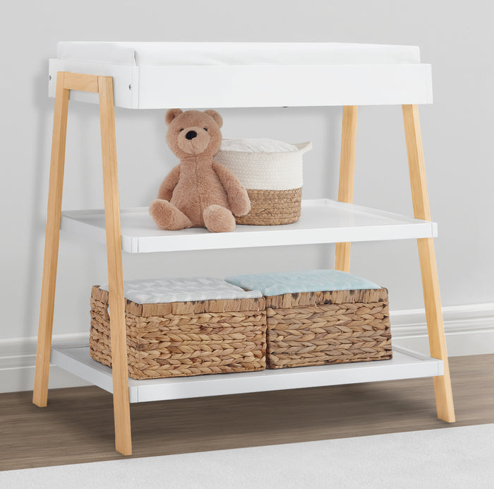 A gray Scout Changing Table with two open shelves, placed in a nursery room corner, with a teddy bear and a wicker basket on one of the shelves. 1-Scout Changing Table