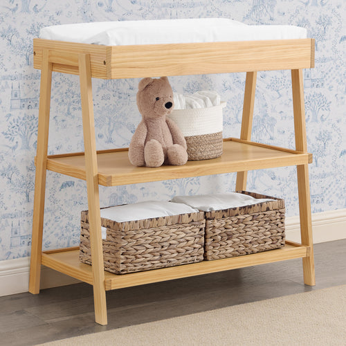 Wooden changing table with storage baskets and a teddy bear against a floral wallpaper background