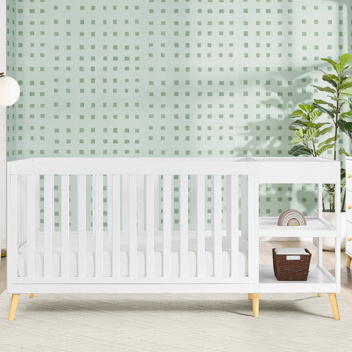 A white Essex 4-in-1 Convertible Crib and Changer with curved design and wooden accents, placed in a room with a green wall and furniture. 2-Essex 4-in-1 Convertible Crib and Changer