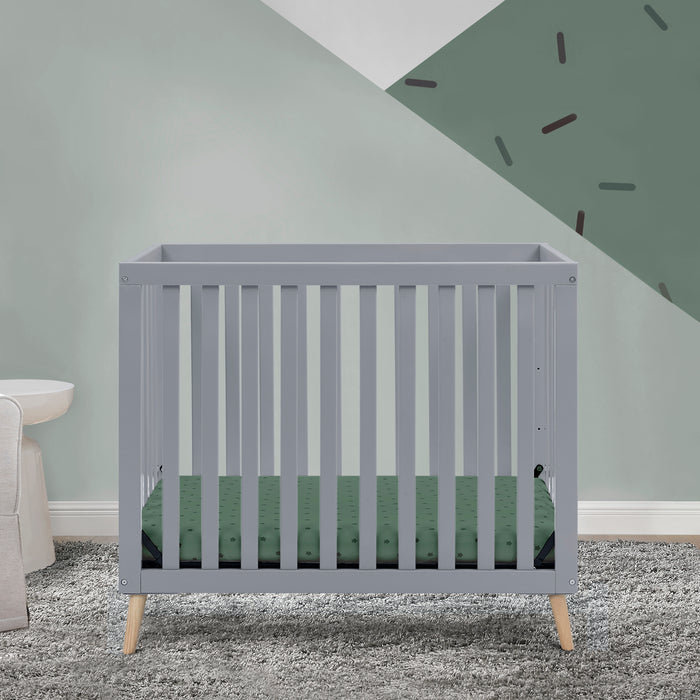 Gray crib with green mattress in a room with gray walls and carpet. 1-Essex Convertible Mini Baby Crib with 2.75-Inch Mattress, Greenguard Gold Certified