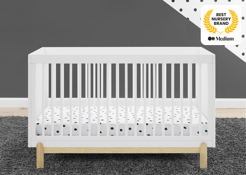 White crib with wooden legs on a gray floor, against a dark gray wall with a 'Best Nursery Brand' award badge. 20-Poppy 4-in-1 Convertible Crib
