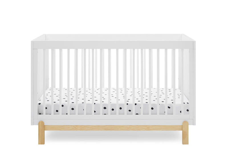 A white wooden convertible crib with a natural oak base and a white mattress featuring a black polka dot pattern, shown against a solid white background.