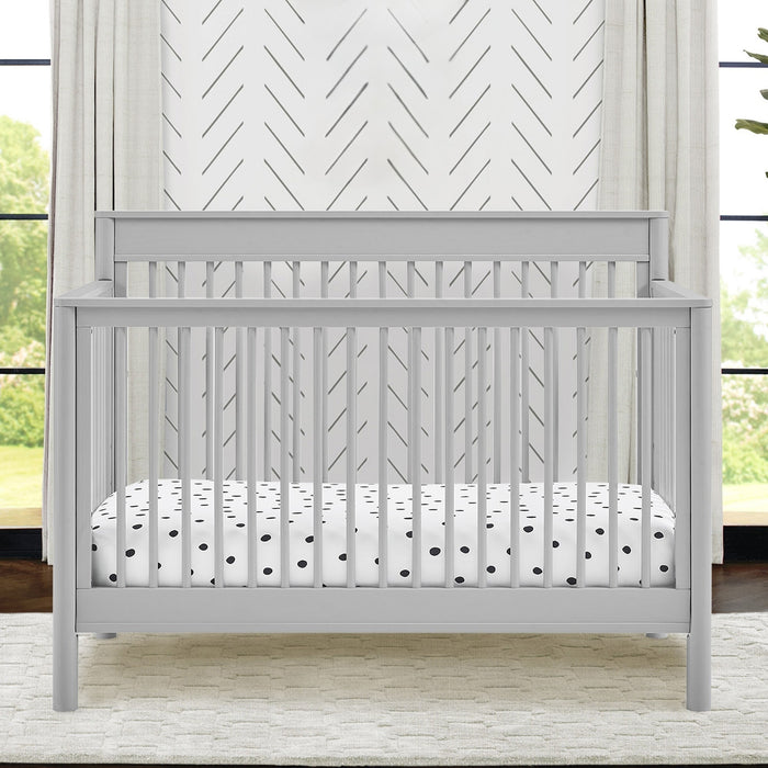 Gray crib with a white mattress featuring black polka dots in a room with large windows. 3-Remy 6-in-1 Convertible Crib