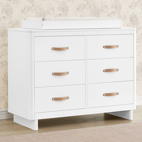 A white 6 drawer dresser with interlocking drawers and brown leather handles, set against a wall with a patterned wallpaper.