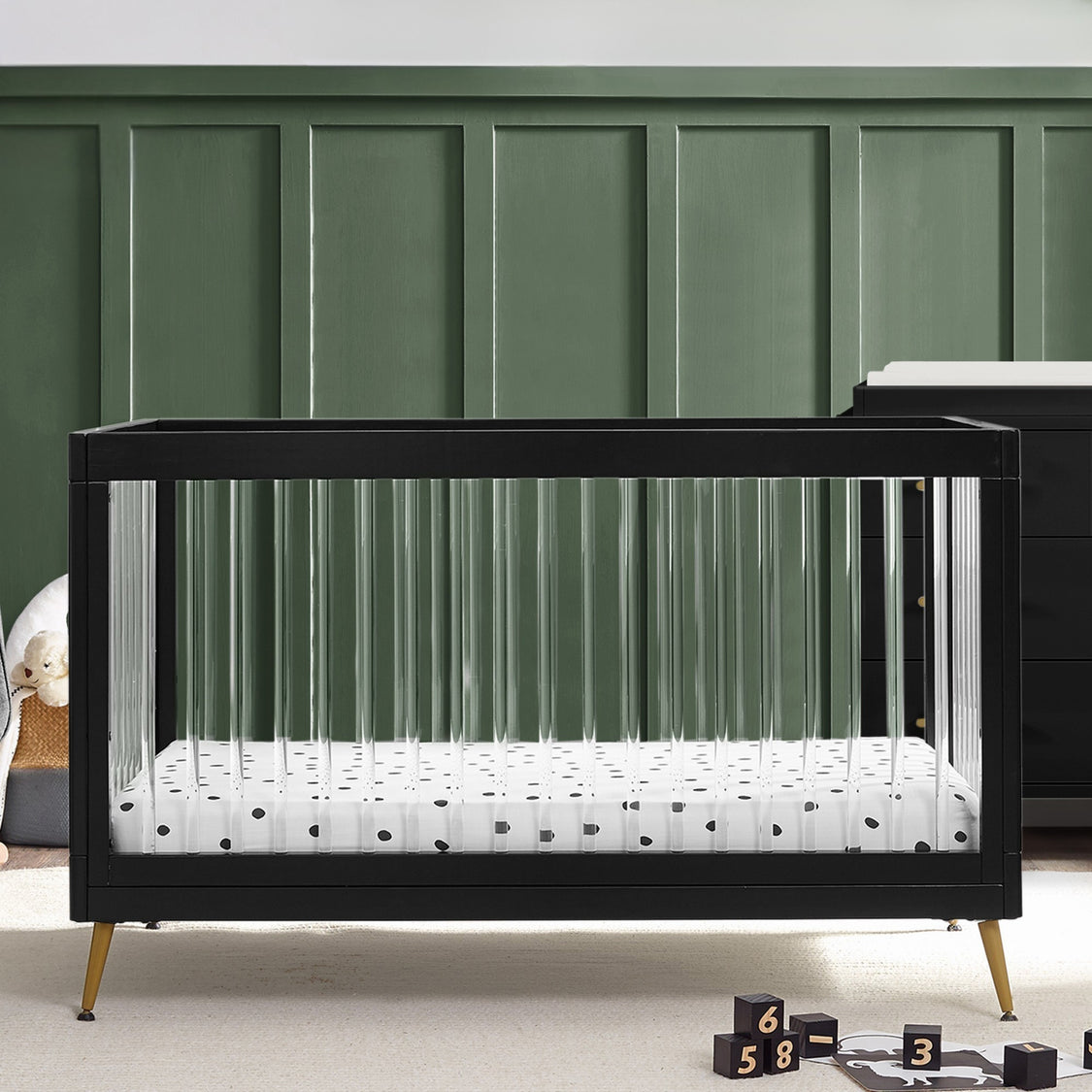 Black crib with white mattress and polka dot sheet against a green paneled wall.