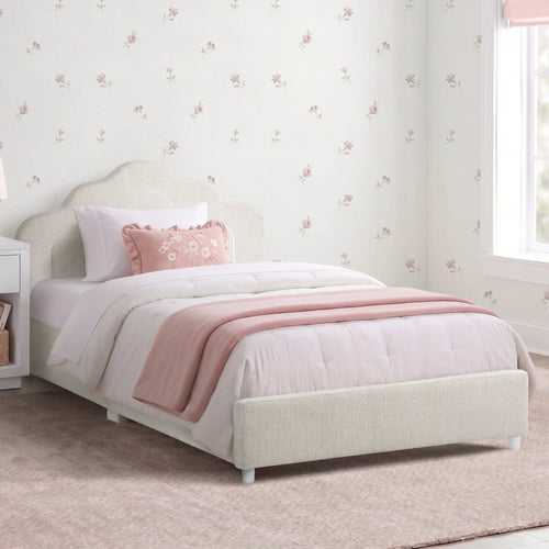 A single bed with a light beige upholstered frame, dressed in white bedding and a dusty pink throw blanket, set in a child's bedroom with floral wallpaper and a window.