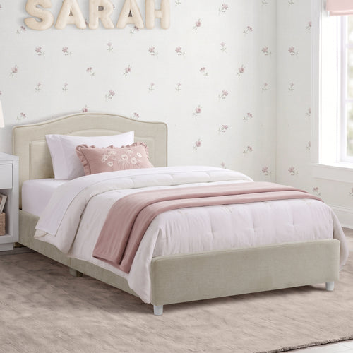A single bed with a light beige upholstered frame, dressed in white bedding and a dusty pink throw blanket, set in a decorated children's bedroom with floral wallpaper and a window.