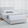 A light gray upholstered single bed frame with a padded headboard and matching footboard, shown dressed in white bedding with a gray accent pillow and folded runner, set against a pale plaid wall. 10-Riley Upholstered Twin Bed