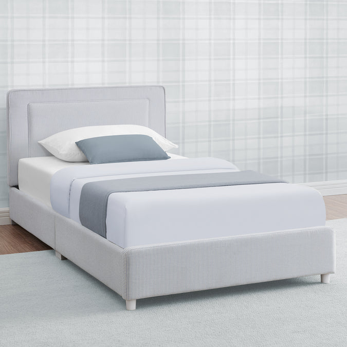 A light gray upholstered single bed frame with a padded headboard and matching footboard, shown dressed in white bedding with a gray accent pillow and folded runner, set against a pale plaid wall. 10-Riley Upholstered Twin Bed
